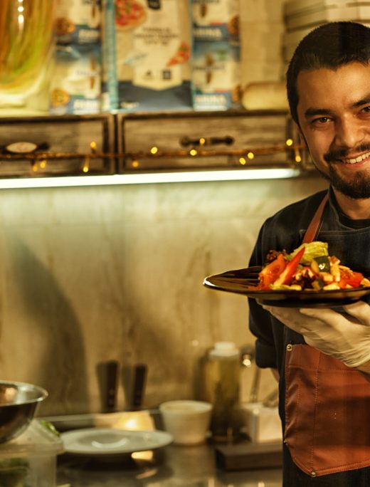Chef smiling and showing delicious salad made of fresh vegetables. Man holding dish with salad in his hand. Background of professional restaurant kitchen with special kitchenware.
